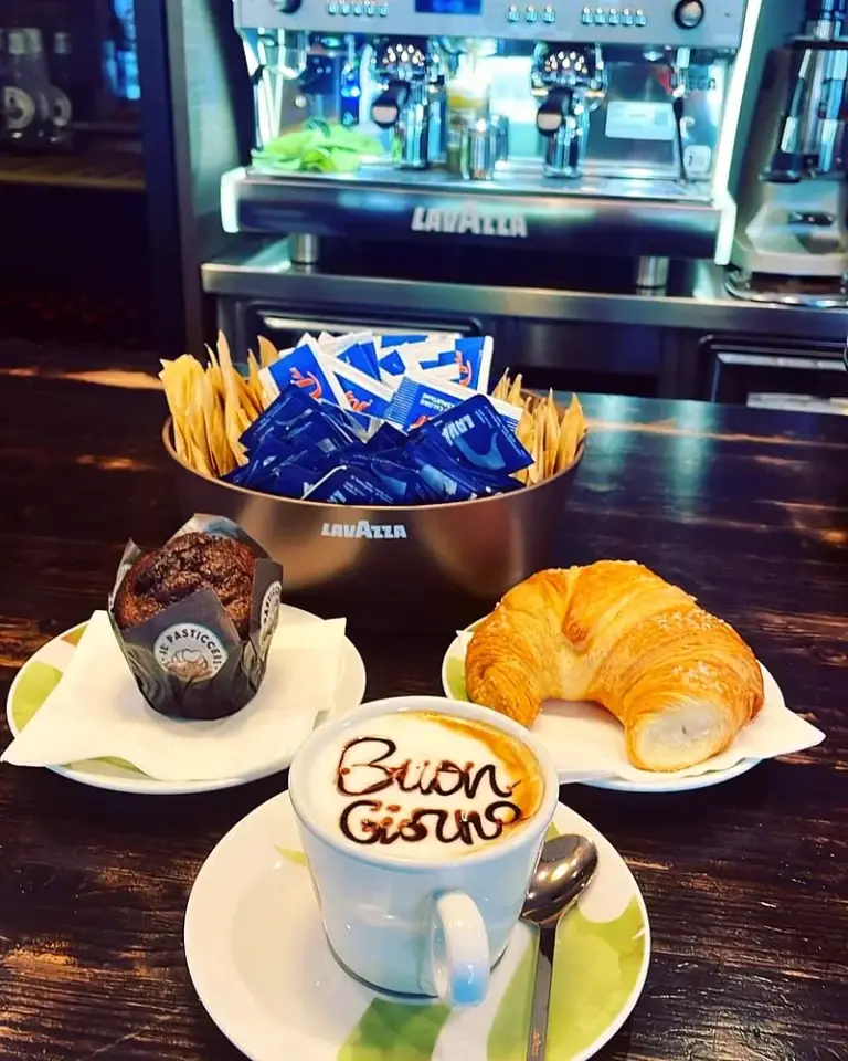 Cappuccino con la scritta "Buon giorno" nella schiuma, un muffin e un cornetto sul banco, con la macchina del caffè dietro
