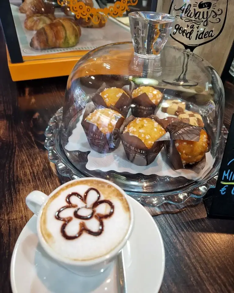Cappuccino con un fiore disegnato nella schiuma davanti alla campana di vetro con i muffin