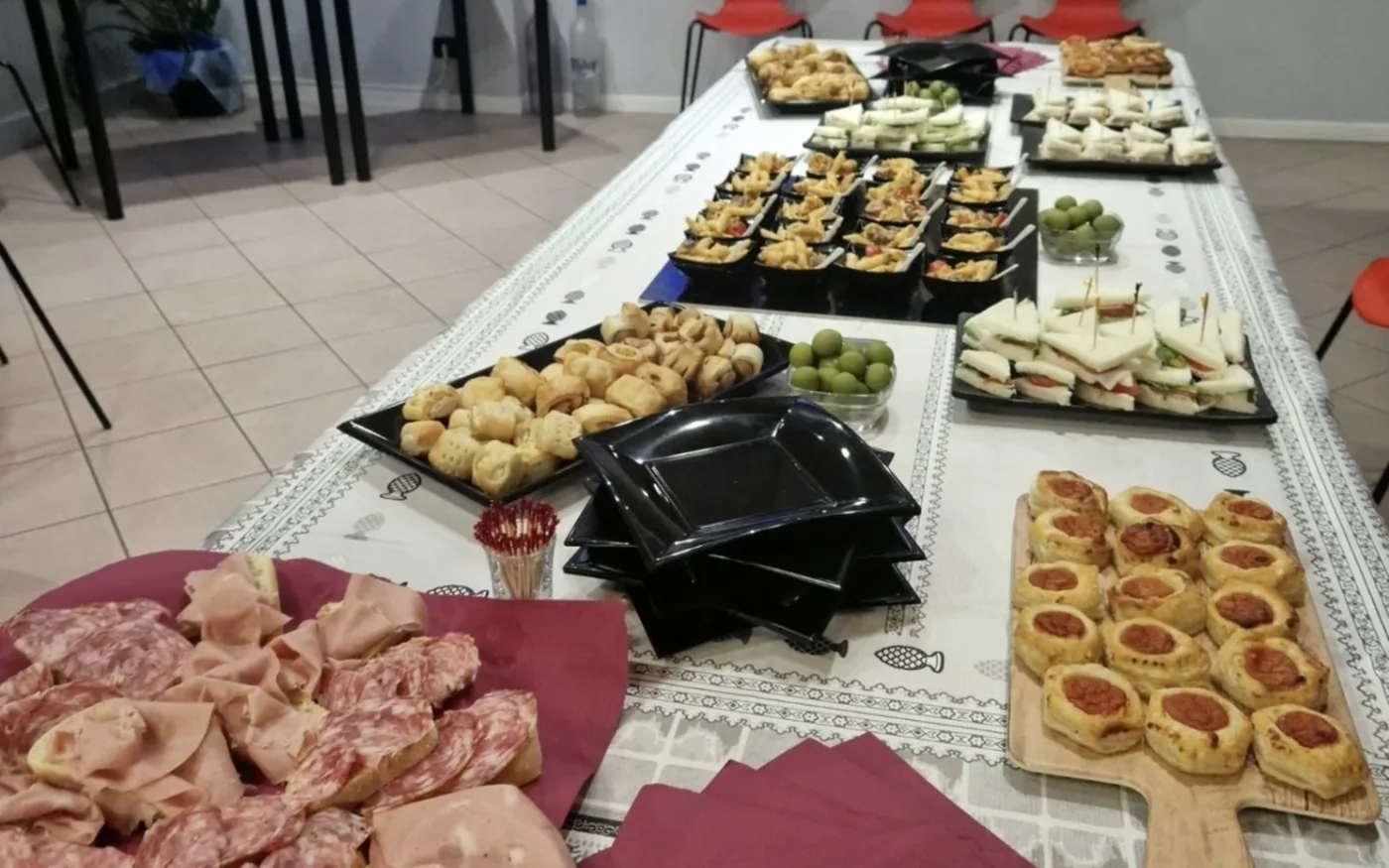 Tavolata lunga apparecchiata con vassoi di rustici, tramezzini, pizzette e affettati, in una sala allestita per un buffet